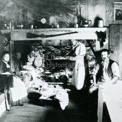 Inside the kitchen of Harmony Vale the home of Jabez Richardson and family taken c1895. Jabez Richardson and his wife Sarah Ann are seated in the chimney ingle. Daughter Martha sits at the end of the table on LHS while daughter Sarah Ann tends the pot. Son John Willie sits smoking his pipe.