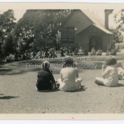 Scene of school children sitting on the ground outside the Mount Dandenong State School building. Perhaps playing a game. Taken in 1938: On back of photograph: Mount Dandenong School 1938
