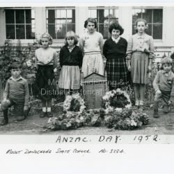 The names of five of the children in the photograph were supplied by Betty Munro after viewing the image on Victorian Collections. L - R: unknown boy, Cynthia Ramage, Betty Munro, Marion Moorby, Frances Barton (nickname 'Bubbles'), Joan Wenborn, unknown boy.: Copy of a photograph showing children from Mt Dandenong State School at an ANZAC Commemoration at the school in 1952. There are four girls and three boys standing behind the School WW1 Honour Board that is surrounded by wreaths of flowers.: On front of photograph: ANZAC DAY 1952 MOUNT DANDENONG STATE SCHOOL NO: 3284