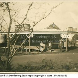Image shows the Mount Dandenong Store and Post Office that replaced the former Post office and Store that was located on the corner of Old Coach and Farndons Roads. The store relocated following the opening of the new Main Road in 1926. The store pictured was on the corner of Helen Road and the Main Road. The image was taken in 1930. The photograph was taken by a member of the Dower family.: Donated as a jpeg digital image that shows the Mount Dandenong Store on the corner of Helen Street and the Main Road in 1930. Two women and a man are standing at the front. Signs advertising the Store & Post Office, Age and Argus are clearly visible on top of the verandah.: Front of image: 1930 Second Mount Dandenong Store replacing original store (Blyths Road)