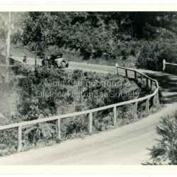 An old fashioned car can be seen pulling away from a bend on the new CRB Road at Wallaby Gully (now Mount Dandenong Tourist Road). Wallaby Gully is near the Mount Dandenong kindergarten. The year is c1926.: Black and white photograph showing a car rounding a bend on a road surrounded by bush and fenced with a wooden rail fence.