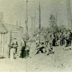The first Mount Dandenong School was established in William Ferguson's shed in 1897. On 11th May 1897 Head Teacher Ethel Gruber opened State School #3284. The locals lined the building and the Education Department supplied the windows. An average of 31 pupils attended. In 1901 the school transferred to a State owned building across the road.: Black and white photograph showing a group of about 17 men sitting and standing beside a simple wooden structure. Handwritten inscription on reverse by John Lundy-Clarke: 1897 Working Bee at the first school on Mount Dandenong which was in Ferguson's shed on Lot 19 Section 2 of the Village Settlement in Blythes Road. Ellis Jeeves is fourth man standing from the right. Ernest Badenhop is thought to be the young man standing side on in centre of picture.
