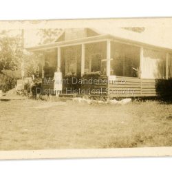 Jim Keogh owned Lot 7 of the Village Settlement. When the new CRB road went through c1920 his block was cut through leaving a small triangle of land on the high side of the CRB road. He built a shop on this piece of land. The store burned down 31/12/1939. The woman on the step in this photo is Win Woodcock.: Sepia image of a woman standing in front of a wooden store. Handwritten inscription on reverse by John Lundy-Clarke: Jim Keogh's shop on the north west corner of Falls and Main Roads. Opposite diagonally from St Michael's Church of England. photo Nov 1926.
