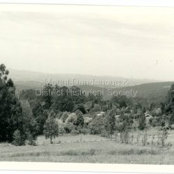 In 1913 Edgar Dower took a photograph from this spot on Ridge Road that became known as Point Clear showing the Mt Dandenong township behind a stand of tall, dead trees. John Lundy-Clarke took this photograph from a similar spot at the Mt Dandenong Arboretum in 1975. Black and white photograph showing a cleared area in the foreground with young tress in the centre and hills in the distance. Several rooftops can be seen mid photo on the right. Reverse of photograph has handwritten description by John Lundy-Clarke.: 1975 View from the Arboretum over Mount Dannong township at the spot where Edgar Dower took the 1913 photo. The Falls gully is in the middle distance. Peter Damman's fence running downhill right foreground.