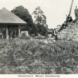 Postcard showing the rock walled base of the Observatory and Trig Point on Mt Dandenong on the RHS and a roofed picnic shelter on the LHS. There are 6 men seated or standing on the rock base.: Front of postcard: Observatory, Mount Dandenong. On back of postcard: Mason, Firth & McCutcheon, Printers, Melbourne.