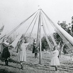 Olinda girls practise the Maypole dance at 'Hillcrest' on Mernda Road in 1911 under the tuition of Vera Devine. From front left circling back are: Lois Lyons, Edie Jackson, Beth Barrow, Phyllis Murray, Amy Allen, Jessie Ebbels, Faith Beatty, Vera Devine, Maggie Schneider, Vera Watson, Hope Barrow, Dolly Witt. The descriptive text on the reverse of this photograph was hand written by John Lundy-Clarke.: 1911 Olinda girls practicing the maypole dance under the direction of Vera Devine at the Devines home Hillcrest Mernda Road. Left to right from front circling 1. Lois Lyons 2. Edie Jackson 3. Beth Barrow 4. Phyllis Murray 5. Amy Allen 6. Jessie Ebbels 7. Faith Beatty 8. Vera Devine 9. Maggie Schneider 10. Vera Watson 11. Hope Barrow 12. Dolly Witt head just showing over ribbon on left. From Mrs A Dodd (Vera Devine) Copied by J.L.C. 1974