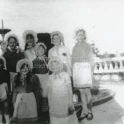 Group of eight young ladies dressed up in bonnets and aprons for a special occasion at Olinda. It may be the Violets Festival. The location appears to be the balcony at Kenloch. Wilma Bartlett, Gwen Evans, Mona Griffin, Norrie Bartlett, Joan Boulter, ? , Jean Patterson, Peggy Hall.