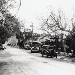 Scene in Olinda township looking up towards range Road. Cars and busses parked along roadside and women and children on footpath. General Store and Gills Newsagency on LHS. c1950.