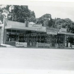 Small photograph from the Olinda township showing view of Southall's Newsagency with milkbar and confectionary. There are many old advertising signs clearly visible. The road is not yet made so this would place the date around the late 1920s.