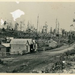 c1908 view of Main Street Olinda with Dodd's store in foreground and Olinda State School up on the hill. Reverse of photograph has handwritten description by John Lundy-Clarke.: Photo of Olinda taken about 1908 showing State School on skyline. Dodds store in foreground. Hodges blacksmith shop on roadside right. Coonara Road, then Mernda Road leads up out the picture right, Mt Dandenong Road goes down left. Range Road (the bullock track) is in immediate foreground and Sassafras turns to the right before the visible tracks on Mernda Road.
