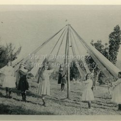 Olinda girls practise the Maypole dance at 'Hillcrest' on Mernda Road in 1911 under the tuition of Vera Devine. From front left circling back are: Lois Lyons, Edie Jackson, Beth Barrow, Phyllis Murray, Amy Allen, Jessie Ebbels, Faith Beatty, Vera Devine, Maggie Schneider, Vera Watson, Hope Barrow, Dolly Witt. The descriptive text on the reverse of this photograph was hand written by John Lundy-Clarke.: 1911 Olinda girls practicing the maypole dance under the direction of Vera Devine at the Devines home Hillcrest Mernda Road. Left to right from front circling 1. Lois Lyons 2. Edie Jackson 3. Beth Barrow 4. Phyllis Murray 5. Amy Allen 6. Jessie Ebbels 7. Faith Beatty 8. Vera Devine 9. Maggie Schneider 10. Vera Watson 11. Hope Barrow 12. Dolly Witt head just showing over ribbon on left. From Mrs A Dodd (Vera Devine) Copied by J.L.C. 1974