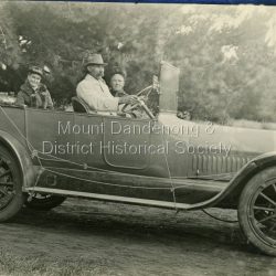 Postcard print showing a Woolrich Service Car with a board attached reading Sassafras & Olinda. George Woolrich started a passenger car service in 1917 with his son Jim. George Woolrich is the driver and his wife Kate Woolrich nee Hand (daughter of early settler Thomas Hand of Kalorama) is seated in the back. The photo would have been taken between 1917 and 1921.