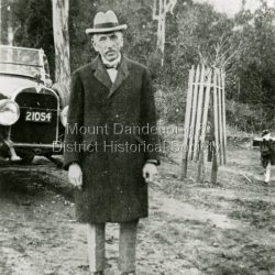 Copy of a photograph of Mr W M (Billy) Hughes, the Australian Prime Minister from 1915 to 1923 in the Main Street of Sassafras. Billy Hughes had a holiday house called Ty-Coed in Clarkmont Road, now number 3A (2025). The Prince of Wales made an unofficial visit in 1920 and stayed with Billy Hughes in his holiday home. The Prince of Wales planted a tree in Anzac Avenue that had been officially opened on April 14th, 1920 by Captain Stanley Melbourne Bruce, who became Prime Minister after Billy Hughes.: Black and white photograph showing well dressed man standing in front of a car .